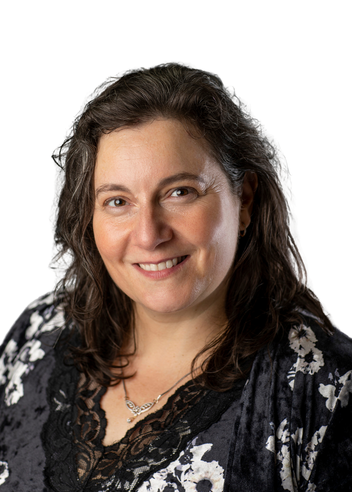 A heat and shoulders picture of a caucasian woman (Zoe) with brown hair and brown eyes smiling at you. she wears a velvet and lace black and white floral blouse