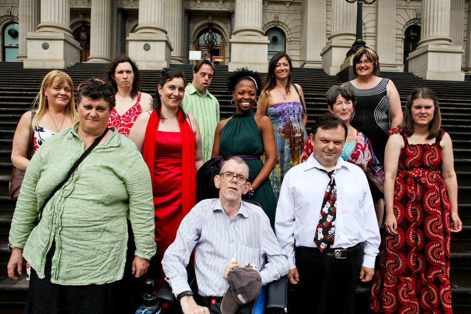 A group of a diverse group of people dressed up people in front of the steps of the Victorian Parliament house. The group includes an older man in a wheel chair. This is the group of people who participated in the whole of person project